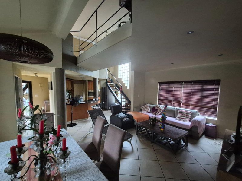 Spacious open-plan living and dining area featuring a double-volume ceiling, mezzanine level, staircase with metal railing, and integrated kitchen access.