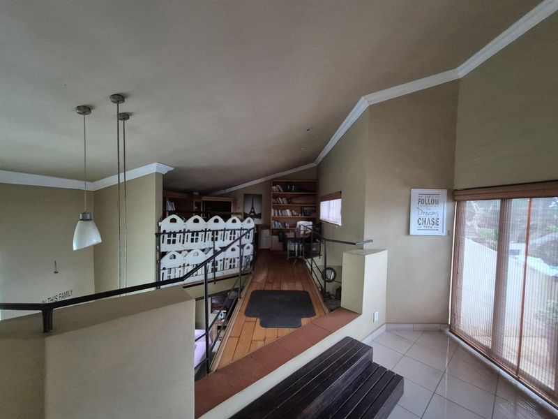 An elevated mezzanine level featuring wooden flooring, a built-in bookshelf, and a metal railing overlooking the lower living area.