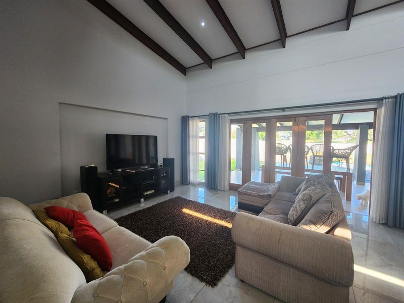 Spacious living room featuring high vaulted ceilings with exposed timber beams, tiled flooring, and large glass sliding doors opening to an outdoor patio area.