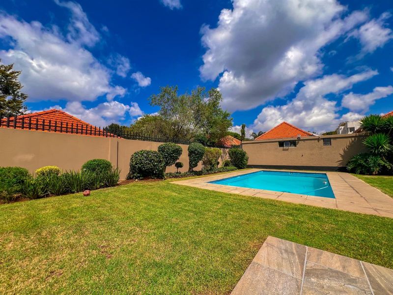 A well-maintained outdoor pool area featuring a clear swimming pool, paved surround, and established garden beds enclosed by a secure perimeter wall.