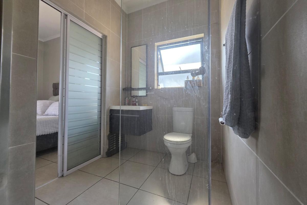 A modern bathroom featuring tiled walls and floors, a wall-mounted vanity, a toilet, and a glass shower screen, with an adjacent bedroom view.