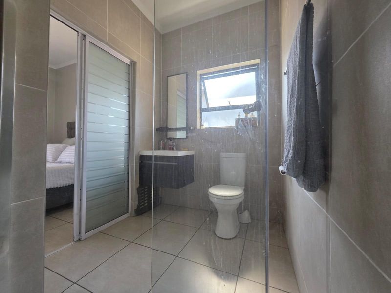 A modern bathroom featuring tiled walls and floors, a wall-mounted vanity, a toilet, and a glass shower screen, with an adjacent bedroom view.