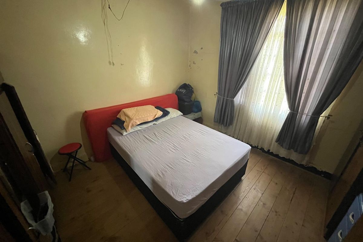 A bedroom featuring timber flooring and a window with curtains. The room requires cosmetic updates to the wall finishes and electrical fixtures.