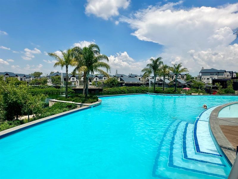 A large, well-maintained communal swimming pool area featuring tiered steps, landscaped surroundings, and views of the surrounding residential estate.