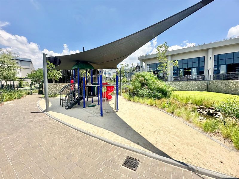 A well-maintained communal playground area featuring a modern shade sail, climbing structure, and slide, set within a landscaped residential complex.