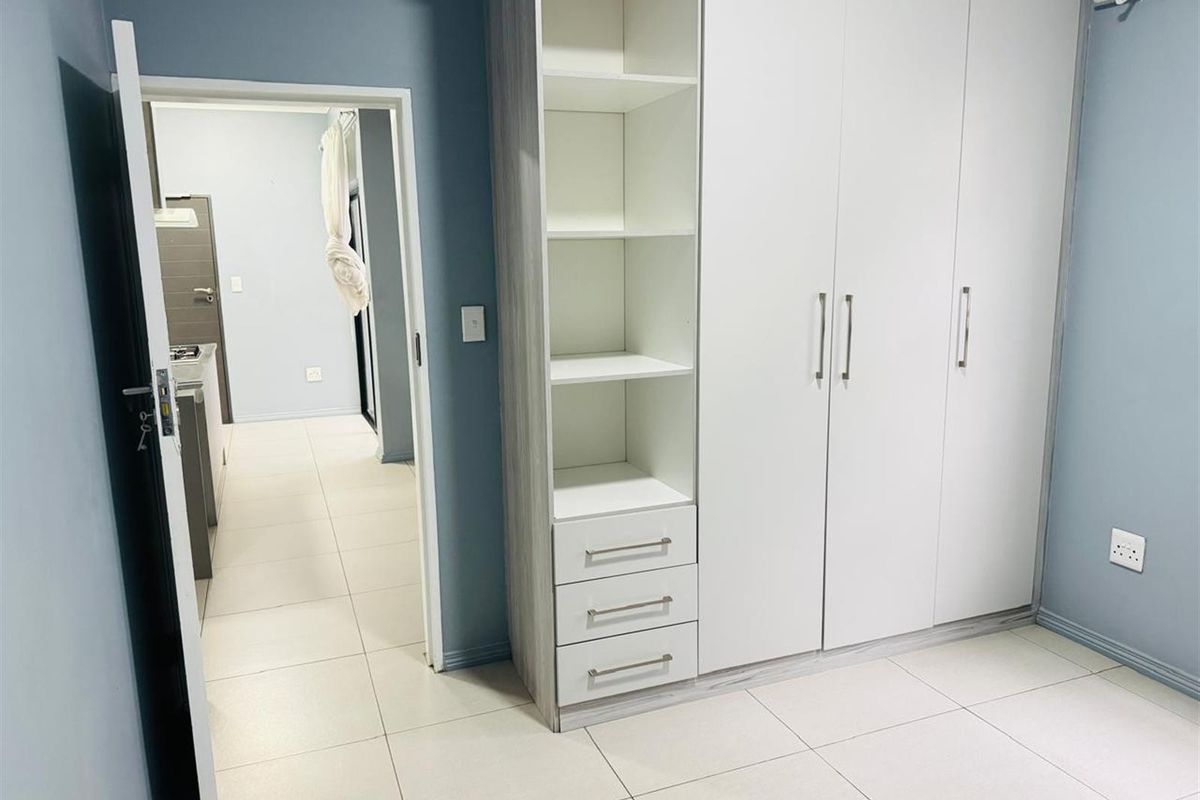 A bedroom featuring built-in cupboards with integrated shelving and drawers, tiled flooring, and a doorway leading to the kitchen area.