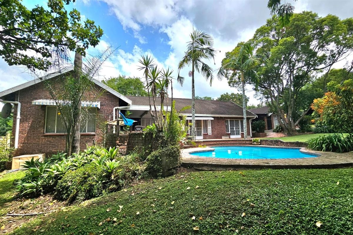 Rear view of a brick residence featuring a swimming pool, established garden, and mature trees in a private outdoor setting.