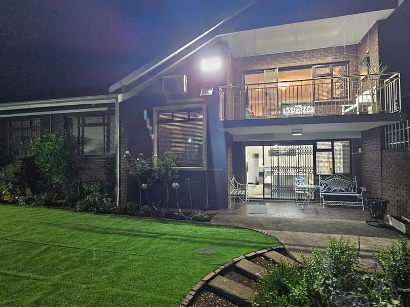 Rear view of a two-story townhouse featuring a brick facade, upper-level balcony, ground-floor patio, and a well-maintained lawn area at night.