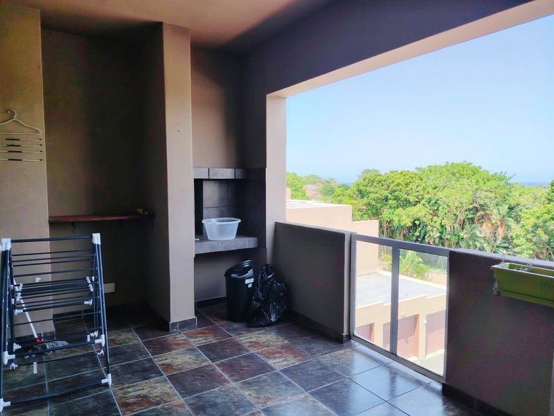 A covered balcony featuring a built-in braai, tiled flooring, and a glass balustrade with views over the surrounding greenery.