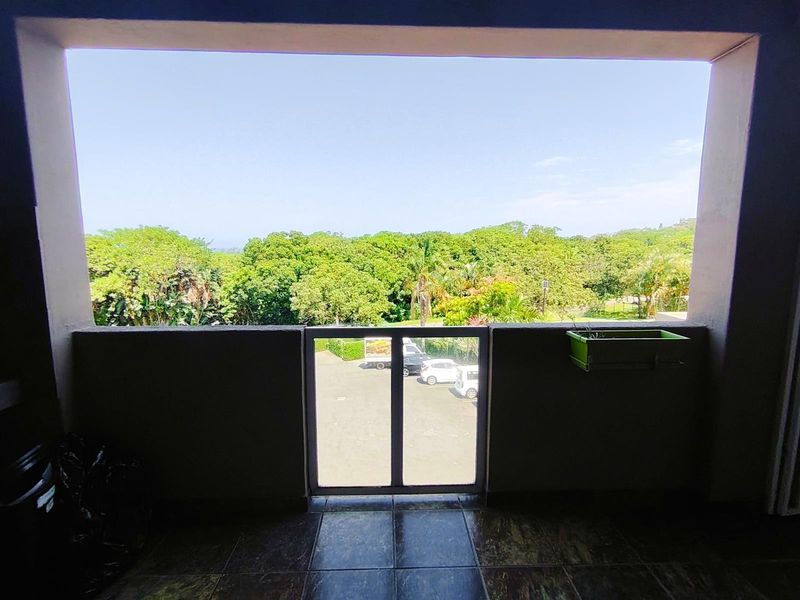A covered balcony featuring tiled flooring and a concrete balustrade, offering an elevated view of the surrounding greenery and parking area.