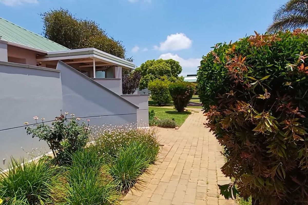A well-maintained exterior rear view featuring a paved walkway leading through a landscaped garden alongside the retirement village unit with a covered patio.