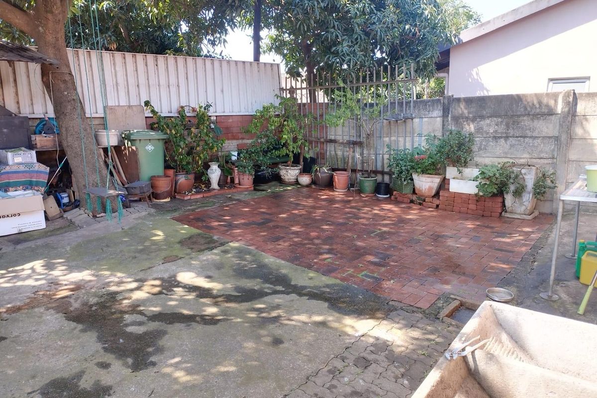A rear outdoor area featuring a mix of concrete and brick paving, enclosed by a boundary wall with mature trees providing shade.