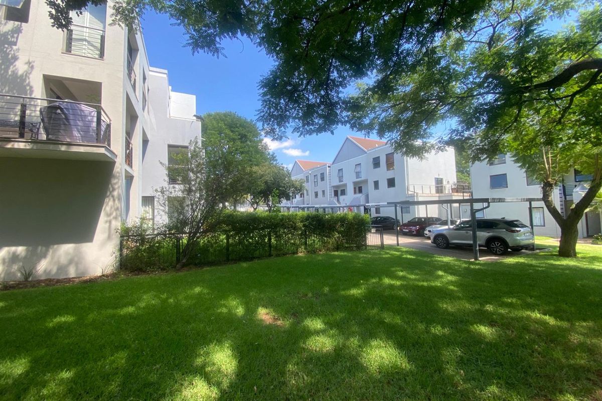 A view of the apartment complex exterior featuring a well-maintained lawn, mature trees, and covered parking bays.