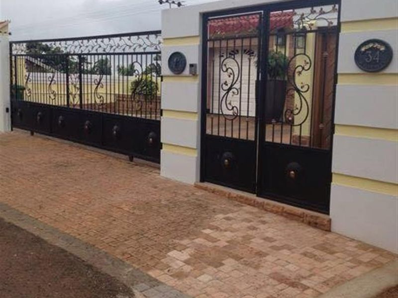 Front entrance featuring a secure wrought iron sliding gate, pedestrian gate, and a well-maintained masonry boundary wall with paved driveway access.
