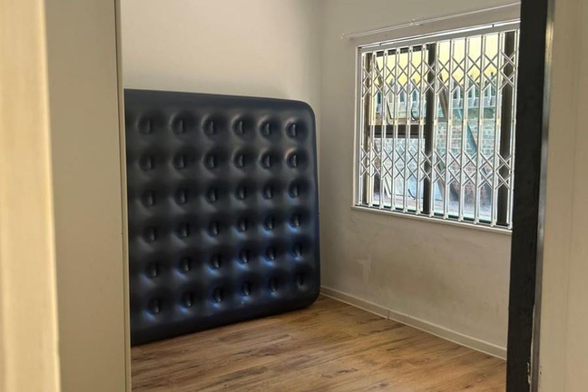 A bright, empty bedroom featuring modern laminate flooring, a ceiling fan, and security bars on the window.