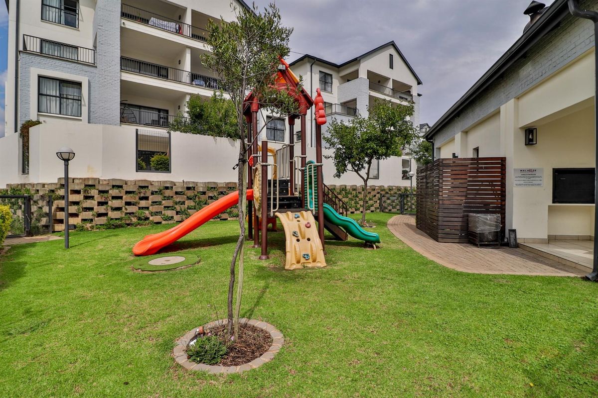 Communal garden area featuring a children's playground, paved pathways, and landscaped retaining walls within an apartment complex setting.