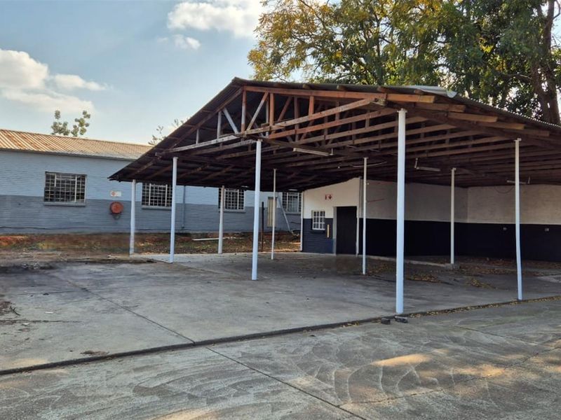 An expansive outdoor covered commercial area featuring a large steel-framed roof structure, concrete flooring, and adjacent brick building facilities.