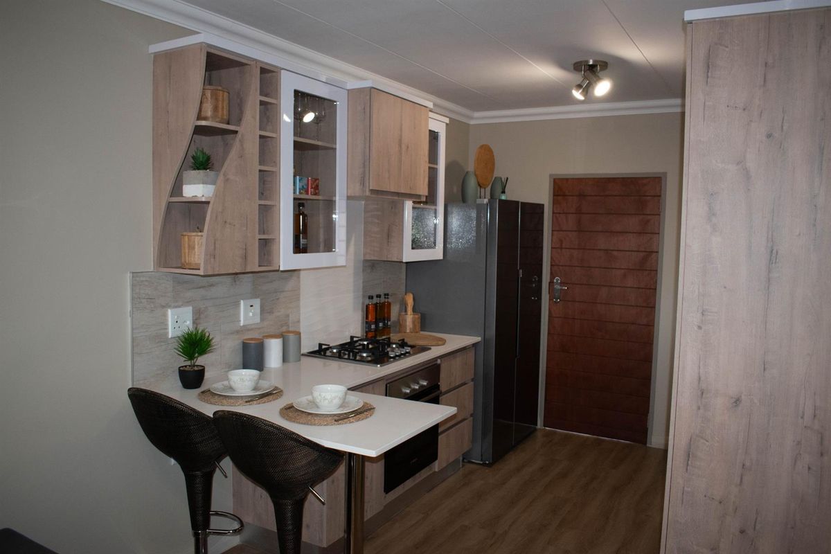 Modern compact kitchen featuring wood-look cabinetry, integrated gas hob, built-in oven, and a breakfast bar extension, finished with neutral tones and laminate flooring.