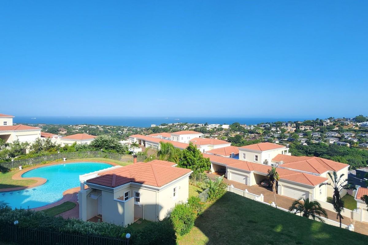 Elevated aerial view overlooking a residential complex featuring a communal swimming pool, tiled roof structures, and a scenic coastal horizon.