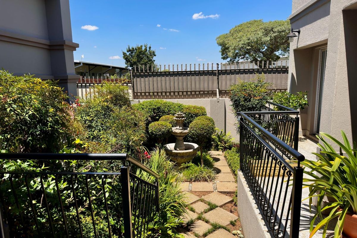 A landscaped rear garden area featuring a stone fountain, paved walkway, and metal security fencing, accessible via a staircase with black metal railings.