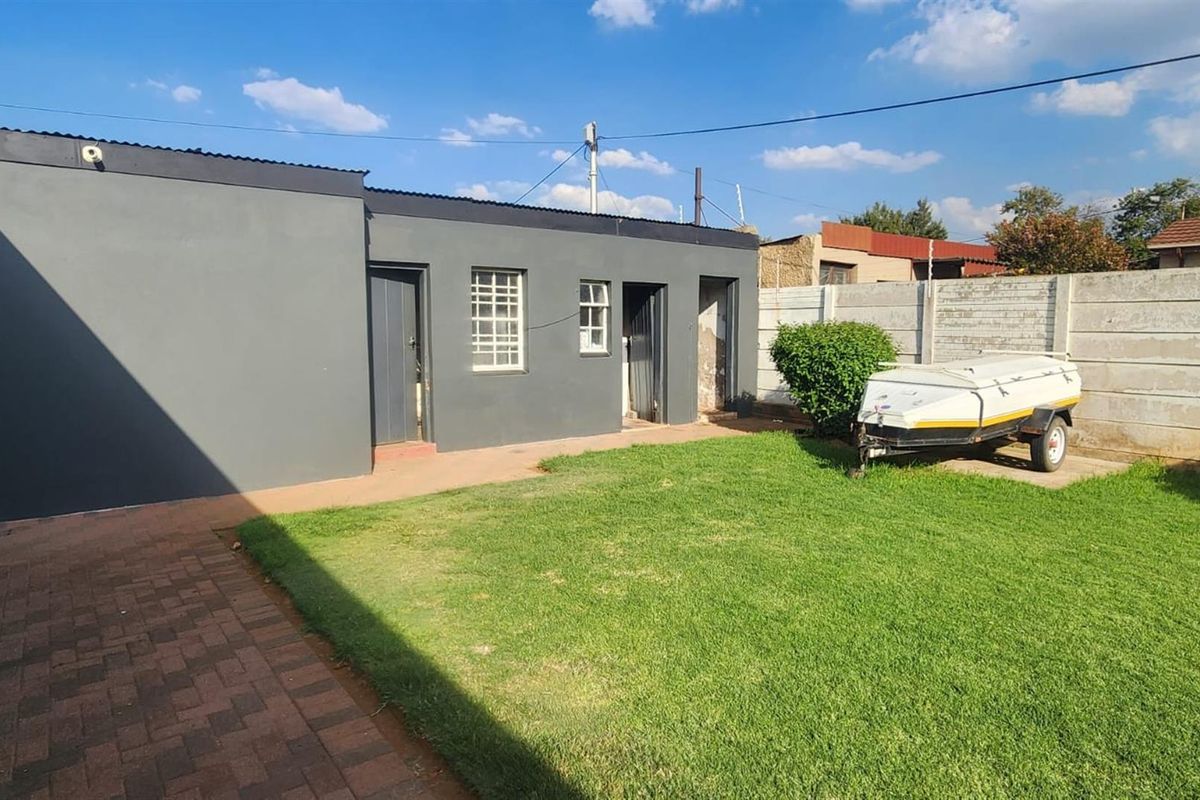 A rear exterior view of the property featuring a paved patio area, a small lawn, and a grey-painted building facade with multiple access points.
