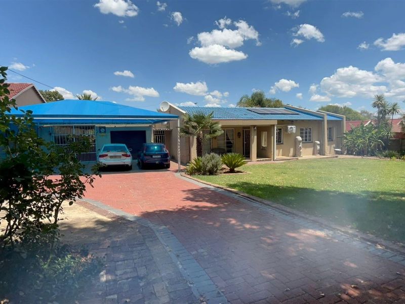 Front view of a single-story house featuring a paved driveway, blue carport, tiled roof with solar panels, and a well-maintained front lawn.
