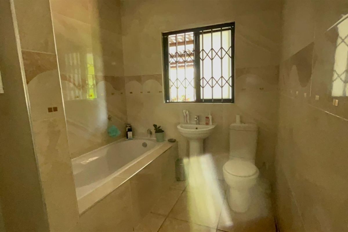 A functional bathroom featuring a built-in bathtub, pedestal basin, and toilet, finished with neutral wall and floor tiling and a security-grilled window.