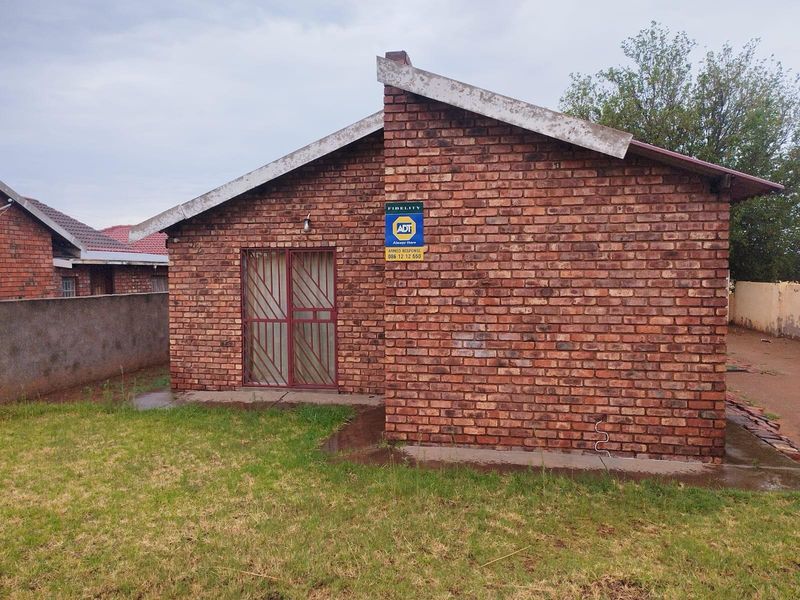 A detached brick-faced house featuring a pitched roof, security gate entrance, and an integrated security alarm system, set within a grassed yard.
