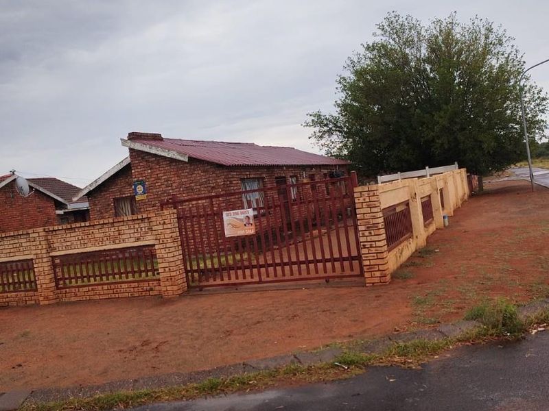 A single-story brick house featuring a metal roof, enclosed by a perimeter brick wall and a metal gate, situated on a corner plot.
