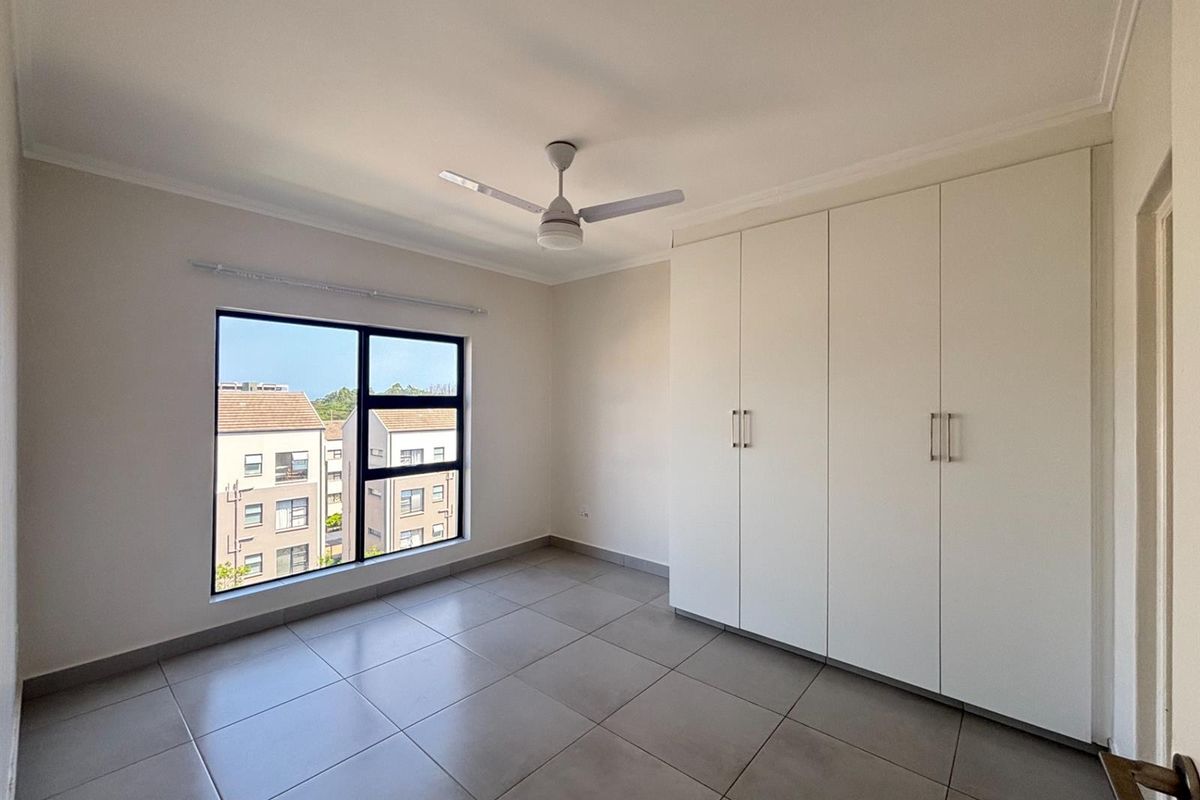 A bright and spacious bedroom featuring neutral floor tiles, built-in cupboards, and a ceiling fan, offering a clean and modern living space.