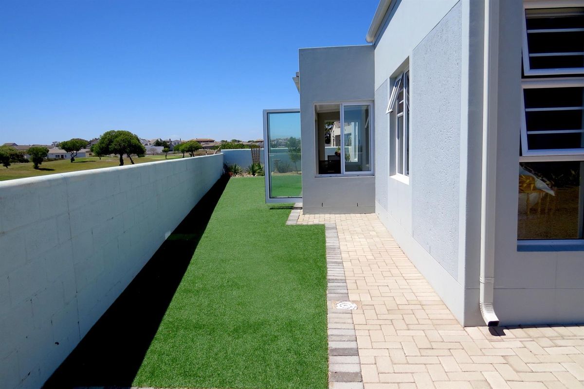 A well-maintained rear exterior area featuring a paved walkway, artificial lawn, and a solid boundary wall with clear views of the surrounding neighborhood.