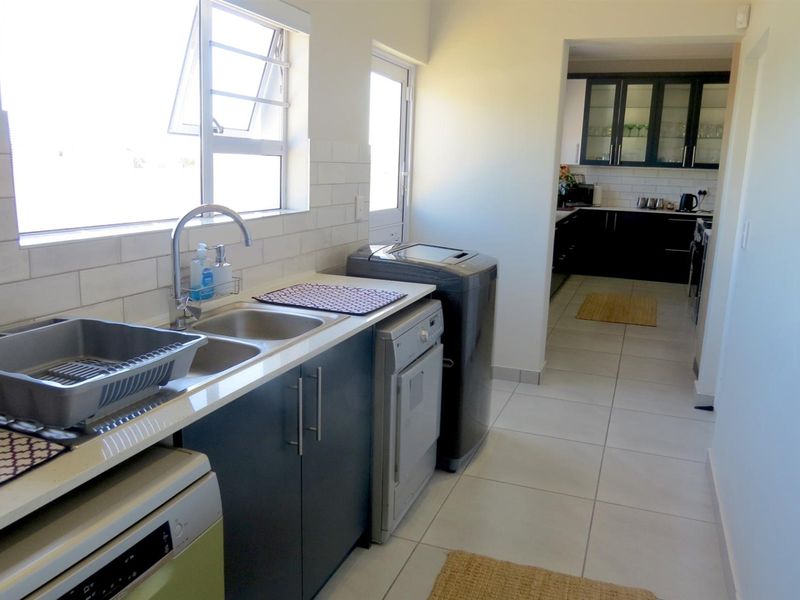 A functional scullery featuring modern dark cabinetry, a double stainless steel sink, subway tile backsplash, and tiled flooring, with an open transition to the kitchen.