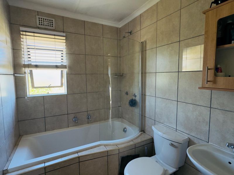 A functional bathroom featuring a bathtub with a glass shower screen, pedestal sink, toilet, and neutral wall tiling.