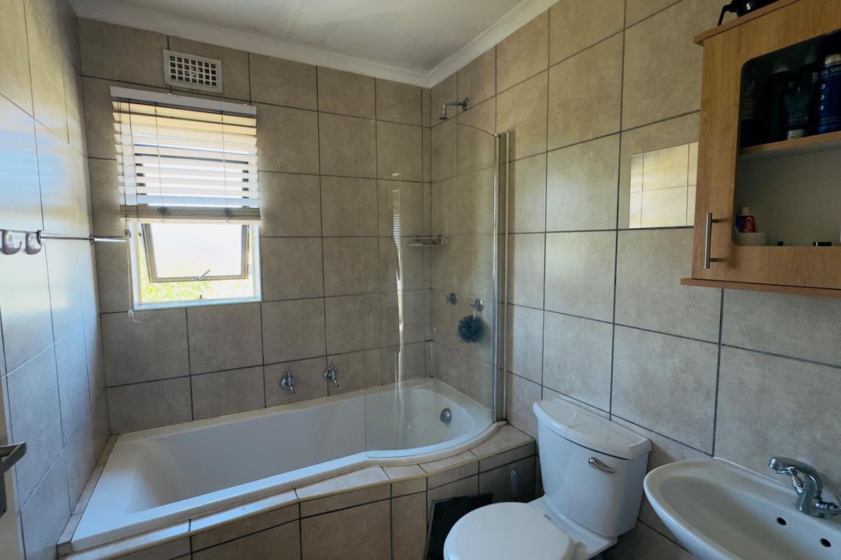 A functional bathroom featuring a bathtub with a glass shower screen, pedestal sink, toilet, and neutral wall tiling.