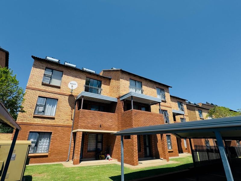 Exterior view of a multi-story face brick apartment complex featuring covered balconies, solar water heating systems, and dedicated carport parking.