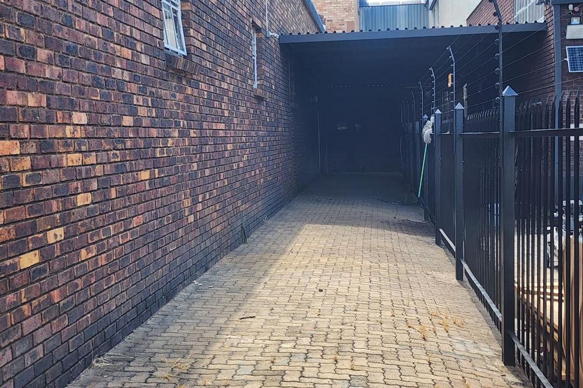 A narrow paved side passage between brick commercial buildings, featuring a covered walkway section and secure perimeter fencing with electric wiring.