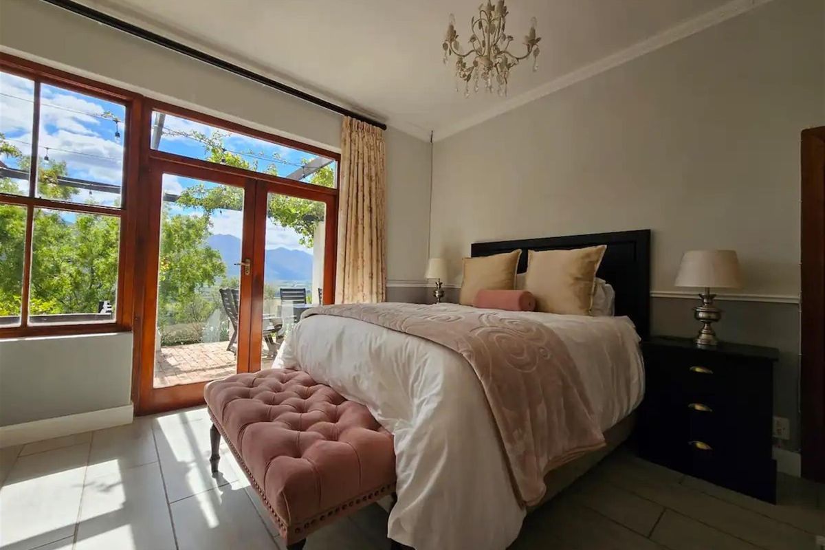 A well-lit bedroom featuring tiled flooring, crown molding, and large wooden-framed glass doors providing direct access to a patio with scenic mountain views.