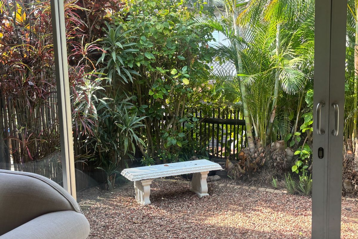 A private rear garden area featuring a gravel base, established tropical landscaping, and a stone bench, viewed through sliding glass doors.
