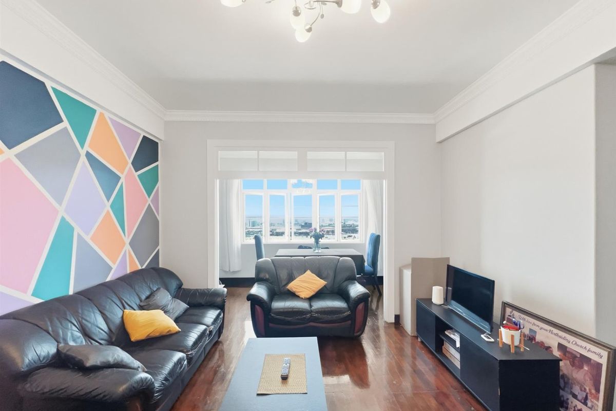 A bright living room featuring polished timber flooring, decorative crown molding, and a connected sunroom area with expansive city views.