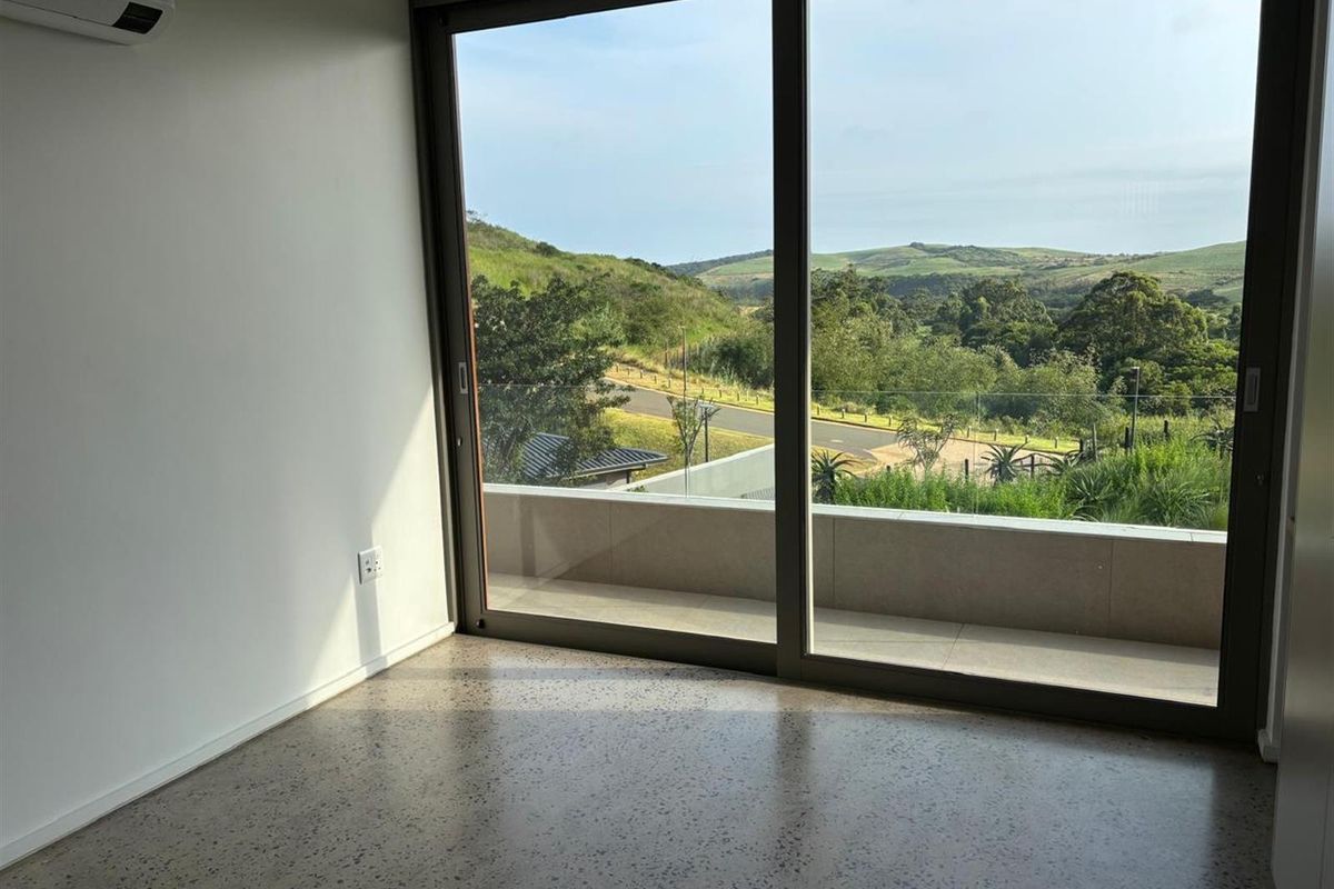 A bright bedroom featuring polished concrete flooring, a wall-mounted air conditioning unit, and large sliding glass doors opening to a balcony with scenic views.
