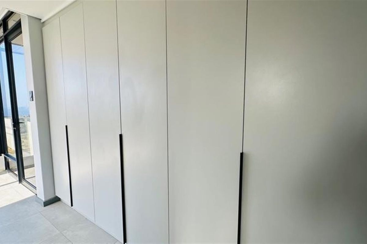 A modern bedroom featuring floor-to-ceiling built-in cabinetry with sleek matte finishes and minimalist handles, adjacent to a glass door leading to a balcony.