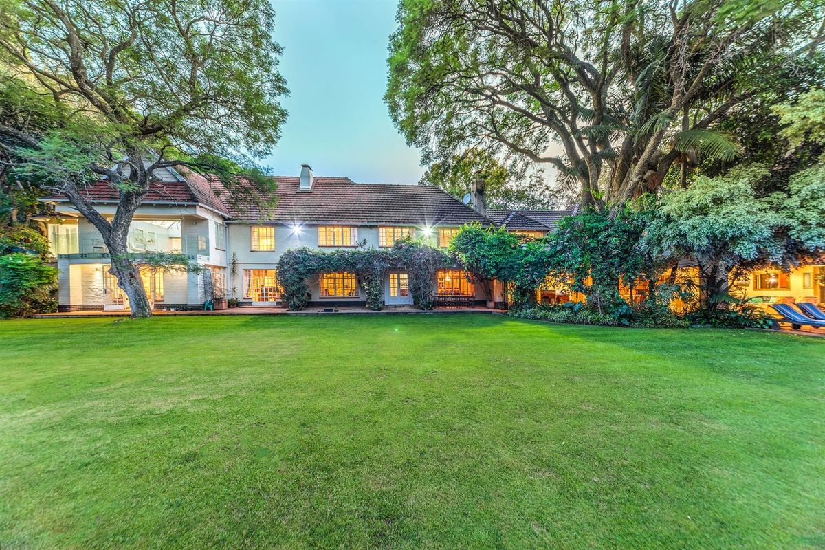 A spacious rear garden view of a large two-story residence featuring mature trees, established landscaping, and a classic tiled roof.
