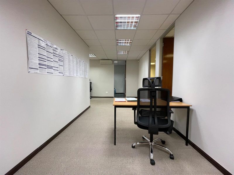 A functional office space featuring carpeted flooring, a suspended ceiling with recessed lighting, and a wall-mounted air conditioning unit.