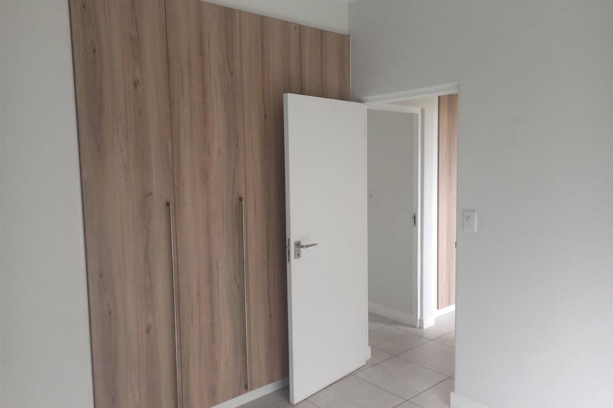 A clean, empty bedroom featuring modern wood-finish built-in cupboards, neutral tiled flooring, and a white internal door leading to an adjacent space.
