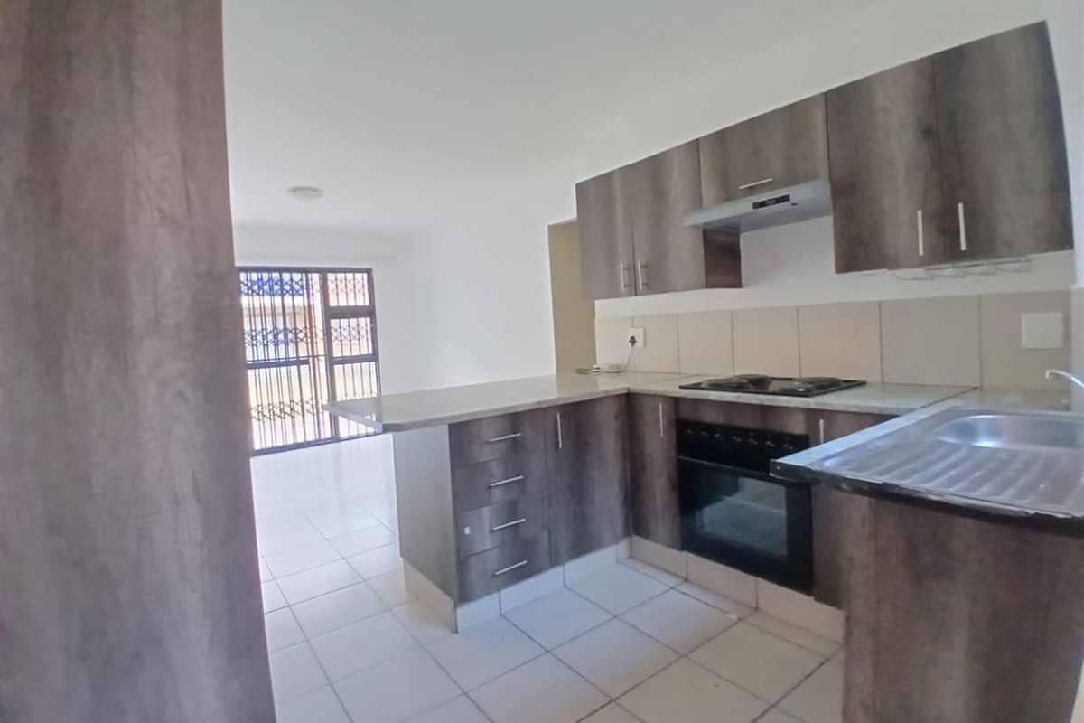 Open-plan kitchen featuring modern wood-finish cabinetry, built-in oven, gas hob, and tiled flooring, flowing into a bright living area with security gate access.