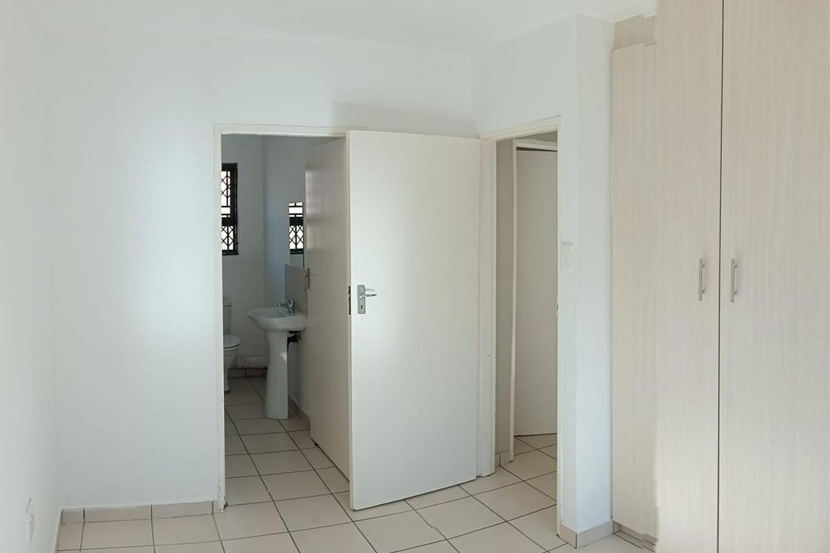 A bright, empty bedroom featuring neutral tiled flooring, built-in cupboards, and direct access to an en-suite bathroom with a pedestal basin and toilet.