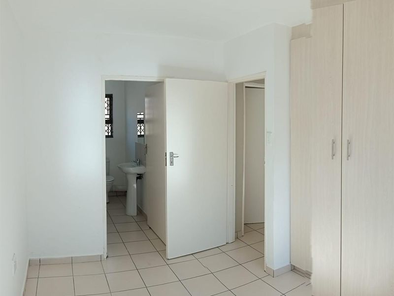 A bright, empty bedroom featuring neutral tiled flooring, built-in cupboards, and direct access to an en-suite bathroom with a pedestal basin and toilet.