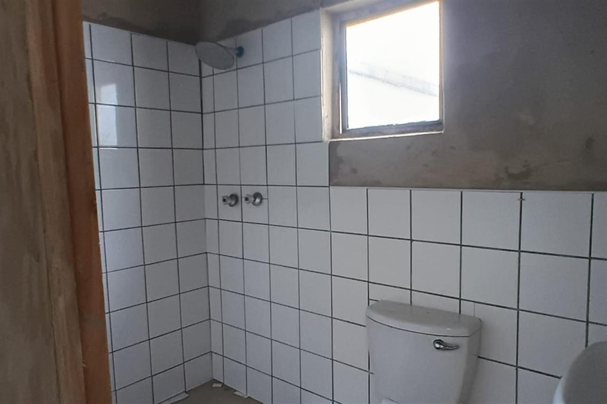 A partially finished bathroom featuring white wall tiles, a shower head, a toilet, and a basin, with unfinished plasterwork visible on the upper walls.