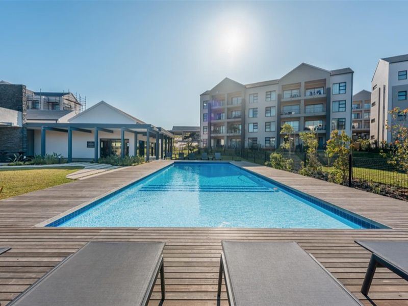A well-maintained communal swimming pool area featuring a timber deck, surrounded by landscaped gardens and modern apartment buildings.