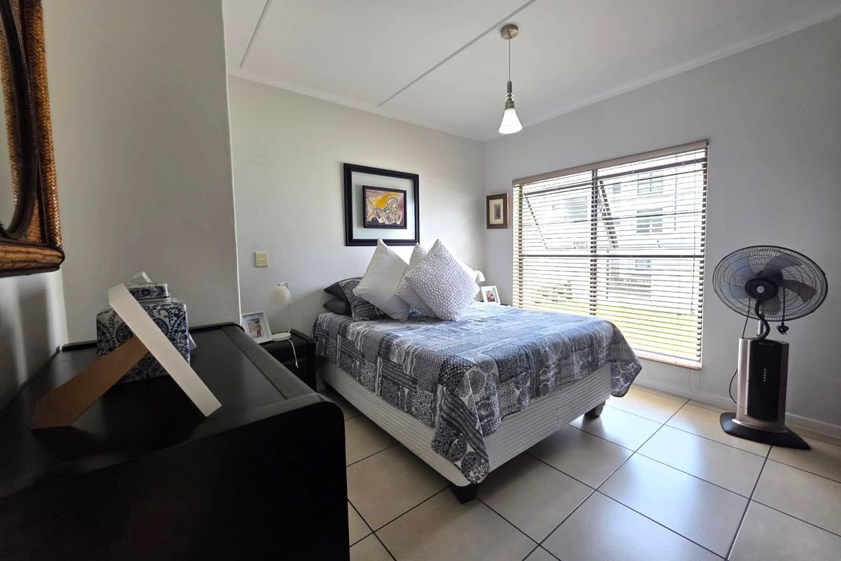 A well-lit bedroom featuring neutral tiled flooring, a large window with fitted blinds, and a simple pendant light fixture.